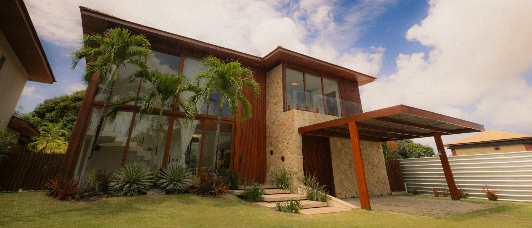 Casa contemporânea de dois pavimentos com fachada em madeira e pedra natural, jardim tropical e palmeiras.