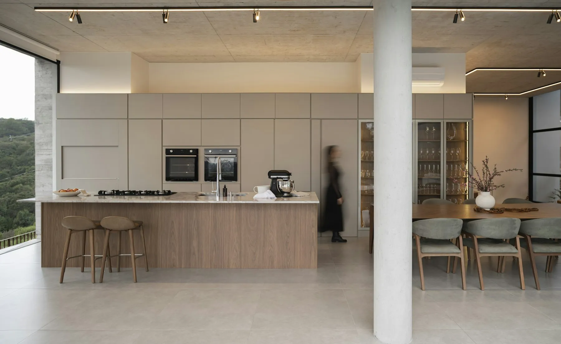 Cozinha integrada à sala de jantar, com ilha em madeira, teto de concreto e grande abertura para a paisagem verde.